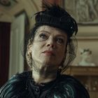 Ich kann den Text nicht erkennen. Eine Frau in viktorianischer Kleidung mit Hut und Schleier schaut nach oben, im Hintergrund ein Raum.