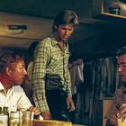 Drei Männer in einem Raum, einer sitzt, zwei stehen. Verschiedene Getränkedosen auf einem Tisch. Rustic Umfeld.