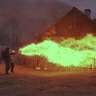 Ein Soldat setzt einen Flammenwerfer ein. Grüne Flammen treffen ein Holzhaus in der Dämmerung.