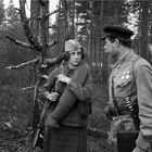 Zwei Personen in militärischer Kleidung stehen im Wald, ein Mann mit Medaillen spricht mit einer Frau, die ein Gewehr trägt.