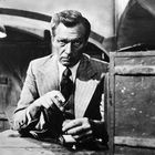 Eine ernste Person in einem Anzug sitzt an einem Tisch, eine alte Werkstatt im Hintergrund. Unten rechts die Zahl "3".