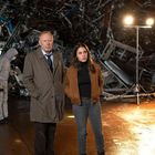 Zwei Personen stehen vor einem Haufen Schrott, beleuchtet von Scheinwerfern, eine Person in Schutzanzug im Hintergrund.