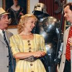 Drei Personen in eleganter Kleidung stehen vor einer Ritterrüstung. Eine Frau lacht. Im Hintergrund hängt ein Gemälde.