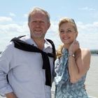 Ein Mann und eine Frau lächeln, stehen vor einem Wasserhintergrund unter einem blauen, leicht bewölkten Himmel.