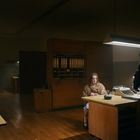 Ein Büro mit Holzmöbeln. Eine Person sitzt, eine steht. Im Hintergrund ein weiterer Mensch bei Schreibtischen. Dämmerige Beleuchtung.