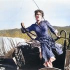 Eine Frau in einem blauen Kleid lenkt energisch einen Wagen in einer ländlichen, bergigen Landschaft.