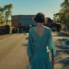 Eine Frau in einem blauen Kleid steht auf einer Straße vor einem umgestürzten LKW, Bäume sind im Hintergrund.