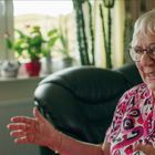 Eine ältere Frau sitzt lächelnd in einem Wohnzimmer mit Pflanzen auf der Fensterbank. Sie trägt eine bunte Bluse. 