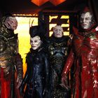 Vier humanoide Wesen mit alienartigen Gesichtszügen in aufwendigen, detaillierten Fantasy-Kostümen in kräftigen Farben.