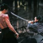 Zwei Personen kämpfen mit Schwertern im Wald, der Mann hat tätowierte und blutige Haut, während die Frau in einer Kampfpose ist.