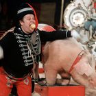 Ein Mann in Zirkusuniform kniet lachend neben einem Schwein. Er hält einen Apfel im Mund und trägt rote Hosen. Im Hintergrund sind Zirkusdekor und Zuschauer erkennbar.