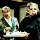 Ein Mann in gemustertem Smoking mit erhobenem Kinn und Schnurrbart. Frau trägt elegantes Kleid, hält Tablett mit Teeservice.