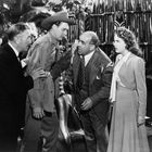 Eine Szene mit vier Personen, die intensiv diskutieren. Ein Mann trägt einen Hut, ein anderer gestikuliert energisch. Pflanzen im Hintergrund.