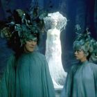 Zwei Personen mit fantasievollen, verworrenen Frisuren stehen nebeneinander. Im Hintergrund erscheint eine leuchtende Gestalt.