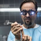 Ein Mann in einem blauen Kittel trägt Lupenbrille mit Lichtern und hält eine Klemme, dabei lächelt er konzentriert.