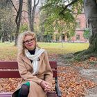 Eine Frau mit Brille sitzt auf einer Parkbank. Herbstlich buntes Laub bedeckt den Boden. Im Hintergrund ein altes Gebäude.