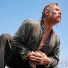 Ein Mann kniet im Freien, hält eine blutende Wunde an seinem Bauch, trägt ein Hemd und schaut in den Himmel.