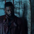 Ein Mann in dunkler Umgebung, trägt eine braune Lederjacke, schaut ernst seitlich, vor einer verwitterten Holzwand.