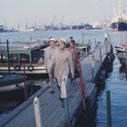 Drei Männer in hellen Anzügen und Hüten laufen auf einem Holzsteg an einem belebten Hafen mit Booten und Kräne im Hintergrund.