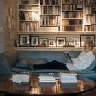 Eine Person liest auf einem Sofa in einer Bibliothek mit vollgestopften Regalen und Fotos an der Wand. Ein Tisch ist vorne.