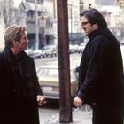 Zwei Männer in dicken Mänteln stehen draußen. Der eine hält ein Stück Papier, während sie sich ernsthaft unterhalten.