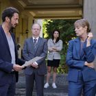 Vier Personen in Businesskleidung stehen diskutierend vor einem Gebäude, eine Person hält ein Blatt Papier.