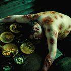 Ein Mann mit Wunden am Rücken liegt über einem Tisch voller leerer Teller und Essensreste, während Ratten darauf kriechen.