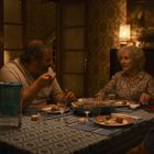 Ein Wohnzimmer mit älteren Tapeten. Drei Personen sitzen an einem Tisch, auf dem gedeckt ist. Warmes Licht erleuchtet den Raum.