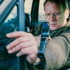 Ein Mann mit Brille zielt konzentriert mit einer Pistole aus einem Autofenster nach draußen.