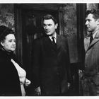 Drei Personen in formeller Kleidung stehen in einem Raum mit gemusterter Tapete.