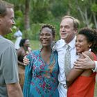 Vier Menschen lachen und umarmen sich in einer grünen Umgebung. Im Hintergrund spielen andere Trommeln.