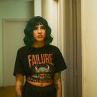 Der Text auf dem T-Shirt lautet „FAILURE“. Eine Person mit dunklem, lockigem Haar steht in einem Flur.