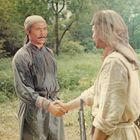 Zwei Männer in traditioneller Kleidung stehen in einer grünen Waldlichtung und schütteln sich die Hände.