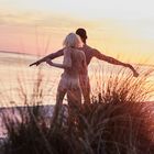 Zwei Personen stehen am Strand, Arme ausgebreitet, im Sonnenuntergang. Hohe Gräser im Vordergrund, Meer im Hintergrund.