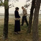 Es gibt keine sichtbaren Texte. Zwei Personen stehen zwischen Bäumen auf einer Wiese. Ein Gebäude ist im Hintergrund zu sehen.