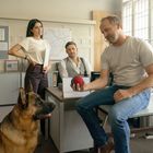 Ein Mann hält einen roten und blauen Ball. Drei Personen und ein Schäferhund sind in einem Büro mit Landkarte an der Wand.