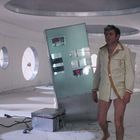 Ein Mann in einem hellen Raum, kreisförmige Fenster, futuristisches Ambiente. Er trägt eine helle Jacke und keine Hose.