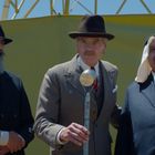 Drei Männer tragen Anzüge, stehen nebeneinander, einer spricht in ein Mikrofon. Im Hintergrund ist ein gelber Bauzaun.