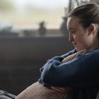 Eine Frau sitzt lächelnd auf einer Couch, umarmt ein Kissen und schaut nachdenklich zur Seite.