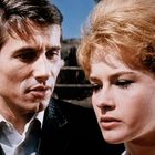 Ein Mann und eine Frau schauen ernst; sie stehen nah beieinander. Im Hintergrund sind Steinstufen zu sehen.