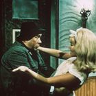 Ein Mann in dunkler Kleidung und Hut hält eine blonde Frau in weißem Kleid in den Armen. Hintergrund: Innenraum mit Holzwänden.