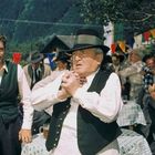 Ein älterer Herr mit Hut und Weste steht im Vordergrund; im Hintergrund sind feiernde Menschen bei einem Fest.