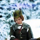 Eine Person in mittelalterlicher Kleidung trägt einen grünen Hut mit einer Feder. Im Hintergrund sind schneebedeckte Bäume.