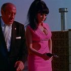 Ein Mann im Anzug und eine Frau in pinkem Kleid halten jeweils eine Waffe, vor einer Ziegelmauer und Schornstein.