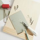 Text "Rico Design" auf einem Linolschnitt-Werkzeug. Werkzeuge und ein Monstera-Blatt-Design auf einer grauen Platte.