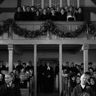 Menschen in traditioneller Kleidung bei einer Versammlung in einer Kirche. Einige stehen auf einer Empore, die mit Girlanden geschmückt ist.