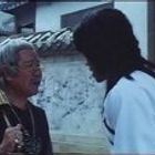 Zwei Personen stehen sich draußen gegenüber, eine trägt ein weißes Gewand, die andere eine dunkle Kleidung und hält einen Gegenstand.