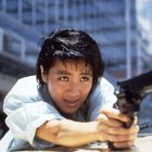 Eine Person zeigt entschlossen mit einer Pistole, über ein Auto gelehnt. Gebäude im Hintergrund.