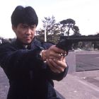 Eine Person im dunklen Mantel hält eine Pistole, angespannt. Hintergrund: Park mit Bäumen und Gebäuden.