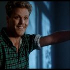 Keine Texte sichtbar. Ein Mann mit lockigem Haar lächelt, trägt kariertes Hemd, lehnt sich an Türrahmen. Licht und Schatten betont.